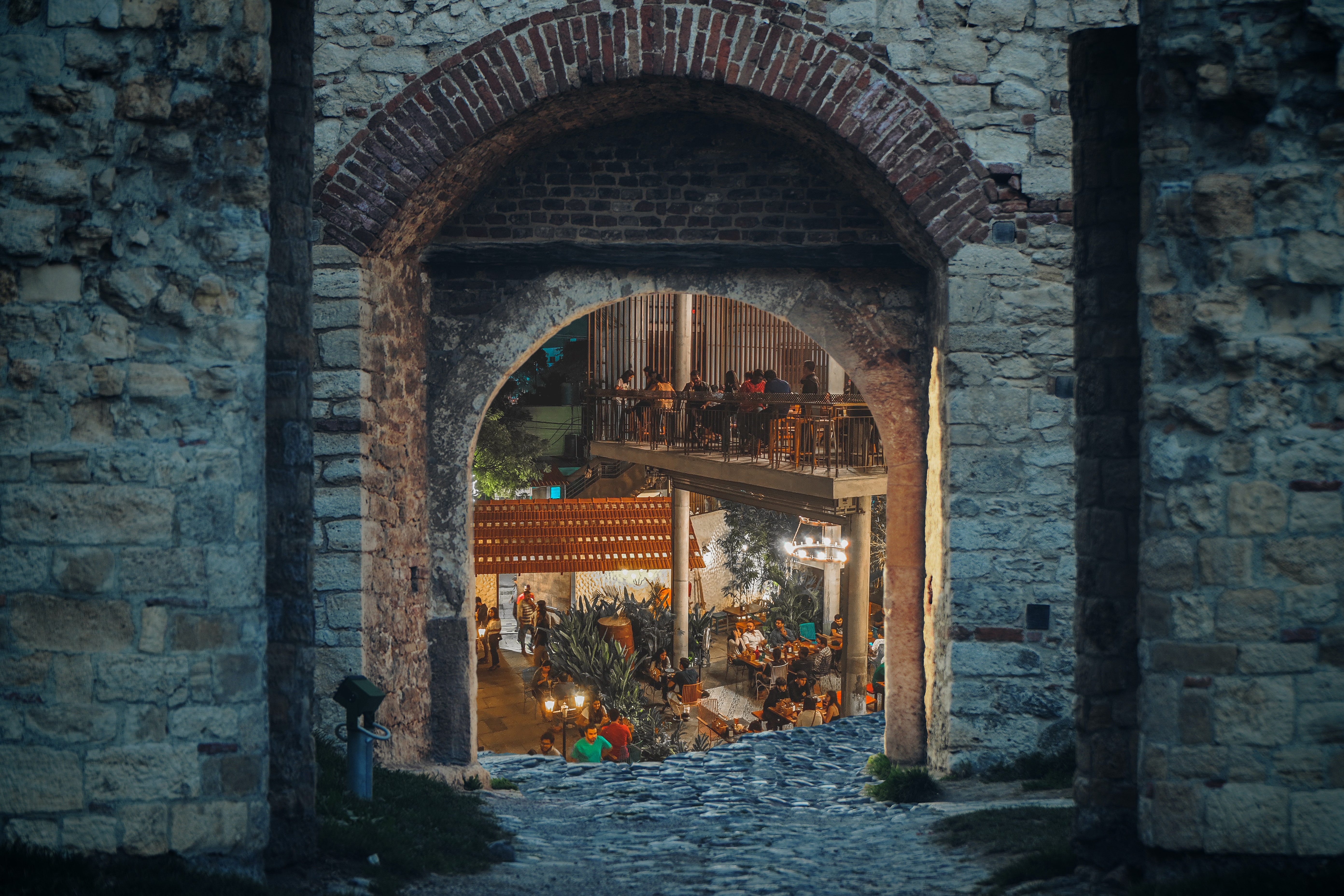 Old stone Greystone fortress walls with an arched reddish brown 2 brick wide soldier course arch over the opening. A smoothed, worn, and well-trodden limestone path leads through the opening, where it splits left and right behind the wall. The background opens to a lower-level courtyard with tables and people gathering in what looks like a celebration.  A second level to the right is supported by two round grey concrete pillars with more people sitting at tables and standing, it is an outdoor courtyard with small trees and red brown tile 